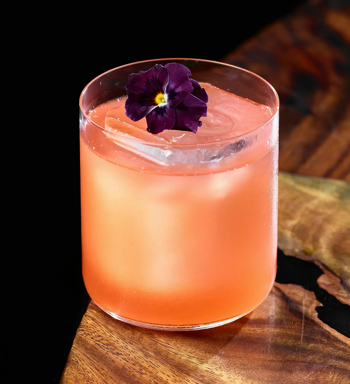 Cocktail color pompelmo con fiore di viola del pensiero su un tavolo in legno.