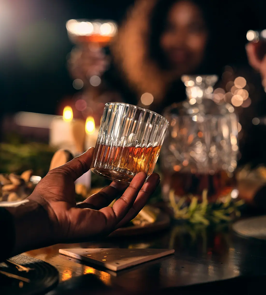 Mano che regge un bicchiere di whisky, persone sfocate sullo sfondo.