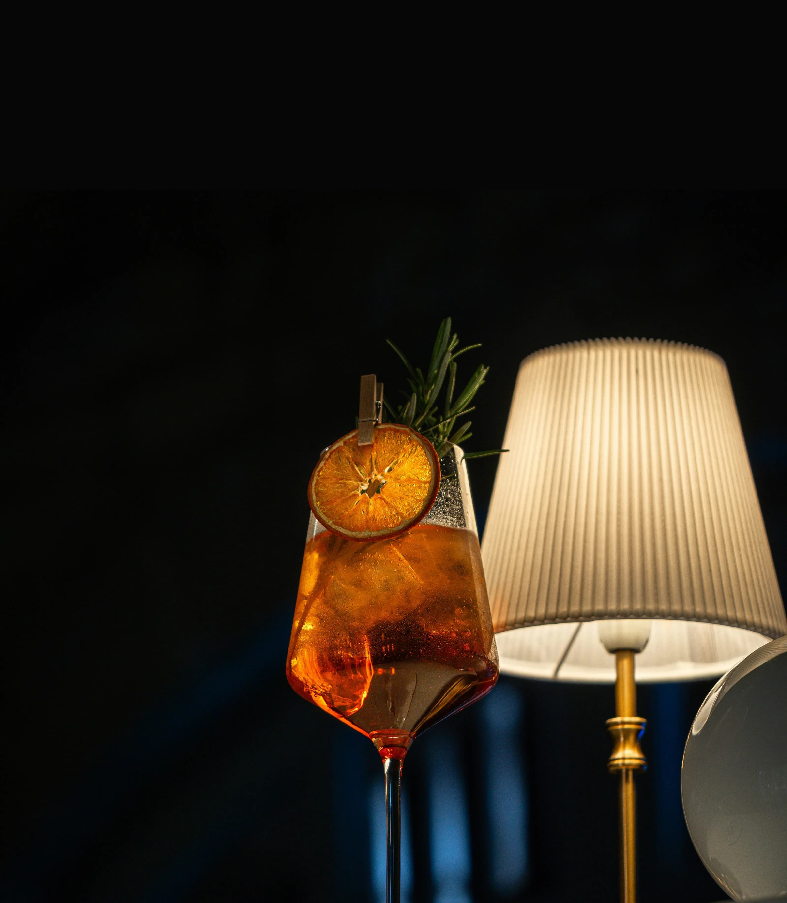 Cocktail ambrato con arancia essiccata e rosmarino, accanto a una lampada calda.